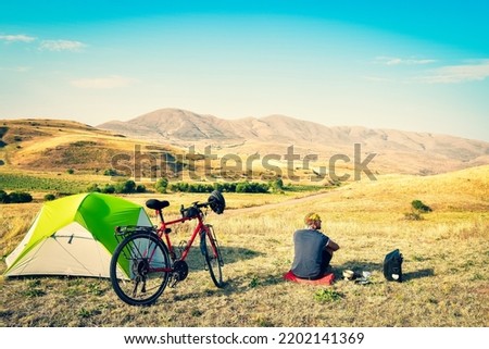Caucasian man sit by touring bicycle in mountains on side of green tent have break time rest in morning. Solo travel journey. Long-term travel around the world