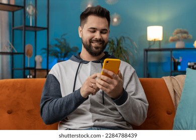 Caucasian man sitting on couch uses mobile phone smiles at home living room apartment. Young guy texting share messages content on smartphone social media applications online, watching relax movie - Powered by Shutterstock