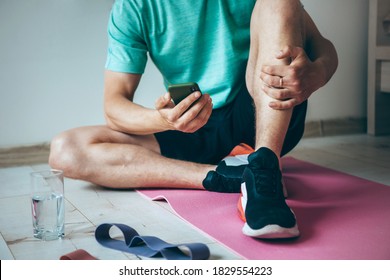 Caucasian Man Sitting On Floor While Relaxing After Doing Home Fitness Is Using A Phone And A Glass Of Water