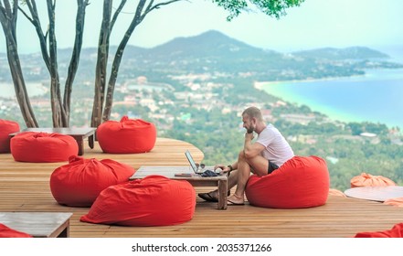 A Caucasian man sitting on a red cushion working remotely with his laptop. There are mountains and the sea in the background - Powered by Shutterstock