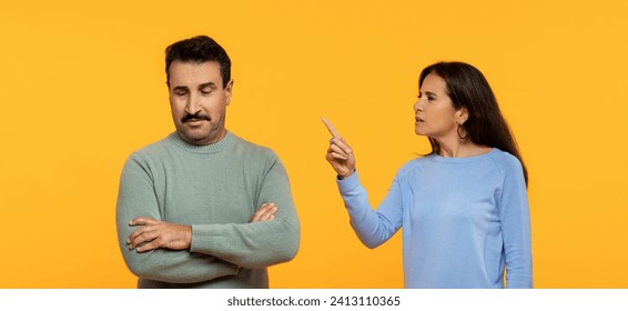 A caucasian man with a skeptical expression crosses his arms while a woman points a finger upwards, suggesting an argument or a discussion, against a bright orange background - Powered by Shutterstock