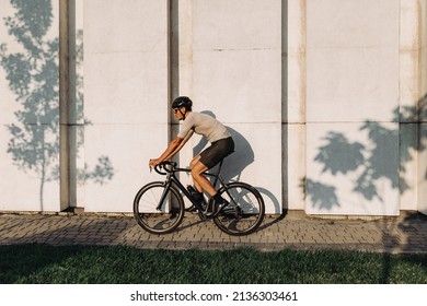 Caucasian Man In Sportswear, Helmet And Glasses Enjoying Outdoors Cycling During Summer Day. Active Guy Riding Bike Near Wall With Shadow From Trees.