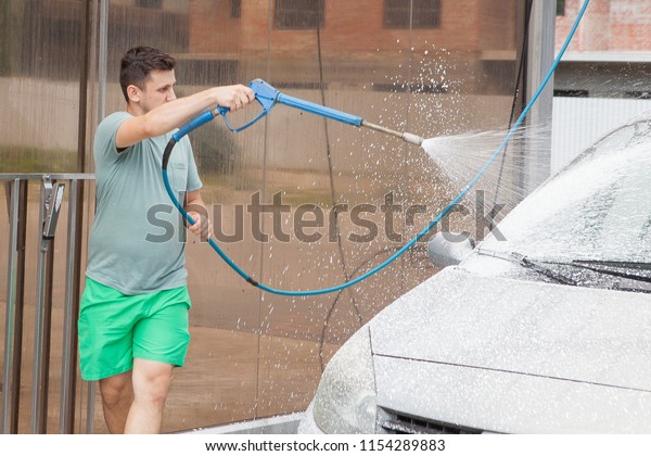Caucasian man is spraying cleaning foam to a\
modern car in car wash with a high water pressure washer. Modern\
car and washing\
outdoors