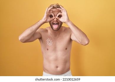 Caucasian Man Standing Shirtless Wearing Sun Screen Doing Ok Gesture Like Binoculars Sticking Tongue Out, Eyes Looking Through Fingers. Crazy Expression. 