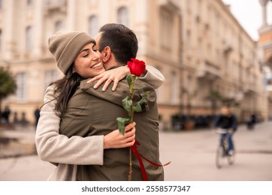 A Caucasian man surprises his female partner with a rose outdoors. She smiles warmly wearing a beanie and coat. The couple is in a city street, reflecting love and joy on International Women's Day. - Powered by Shutterstock