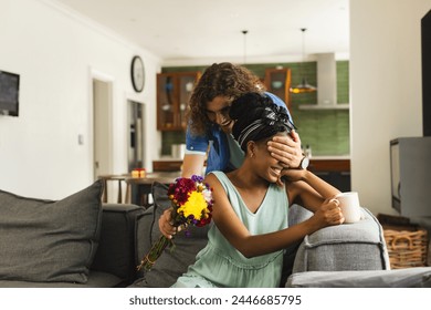 Caucasian man surprising African American woman with flowers at home on couch. The man, with curly brown hair, holds a bouquet to delight his partner, who has a joyful expression, unaltered. - Powered by Shutterstock