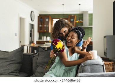 Caucasian man surprising African American woman with flowers at home on couch. The African American woman, with a radiant smile, receives flowers from her Caucasian partner, unaltered - Powered by Shutterstock