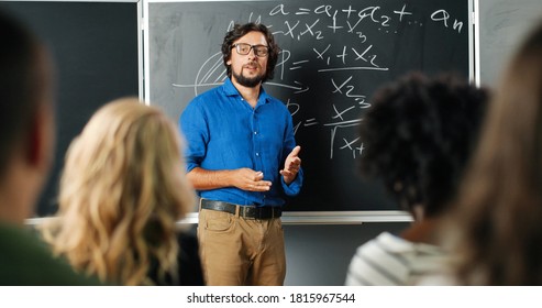 Caucasian man teacher at school at blackboard talking with pupils or students and asking question. Math class concept. Male lecturer in glasses explaining mathematics laws to kids. Educational concept