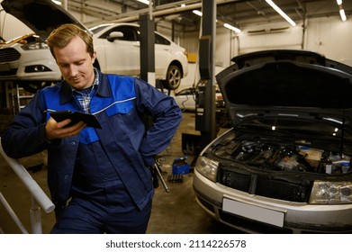 Caucasian Man, Technician, Auto Mechanic In Work Overalls Holding A Wrench In His Hand And Using A Mobile Phone While Standing Against A Car With An Open Hood While Working In A Car Workshop