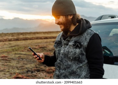 Caucasian man, traveler and adventurer with a beard and tattoos consulting his mobile phone leaning on his 4x4 car at sunset.