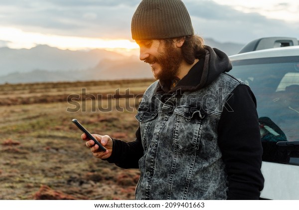 Caucasian man, traveler and adventurer with a\
beard and tattoos consulting his mobile phone leaning on his 4x4\
car at sunset.
