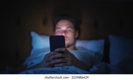 Caucasian Man Uses Smartphone in Bed at Home at Night. Handsome Guy Browsing Social Media, Reading News, Doing Online Shopping, Chatting with Friends Late at Night. Focus on Hand Holding Mobile Phone