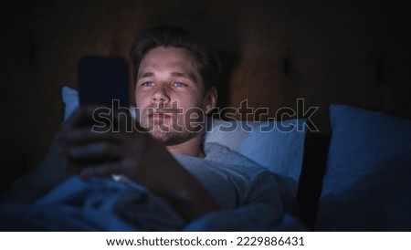 Caucasian Man Uses Smartphone in Bed at Home at Night. Handsome Guy Browsing Social Media, Reading News, Doing Online Shopping, Chatting with Friends Late at Night.