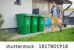 Caucasian Man is Walking Outside His House in Order to Take Out Two Plastic Bags of Trash. One Garbage Bag is Sorted as Biological Food Waste, Other is Recyclable Bottles Garbage Bin.