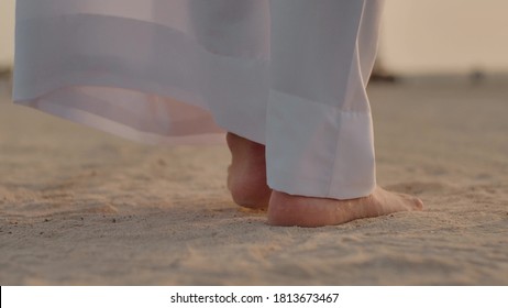 Caucasian Man In White Long Clothes To The Heel, Walking Along The Sand With Slow Steps, Taking His Feet Off The Ground, You Can See The Feet. Slow Step. Back View. Close-up.