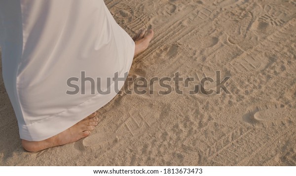 Caucasian\
man in white, long clothes up to the heels walking along the sand\
in small steps. Bare feet walking along the sand. On the sand other\
footprints from the wheels and wheels of\
cars.