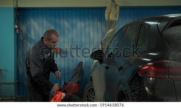A Caucasian man works in a car wash service. A\
man in a worker\'s uniform washes the car thoroughly by spraying it\
with a water pistol.
