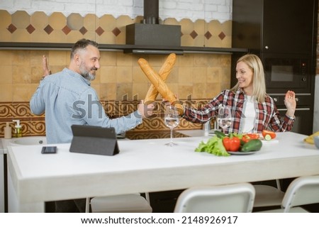 Caucasian married couple in casual attire having fun on kitchen while fighting with freshly baked baguettes. Mature man and woman enjoying carefree moments at home.