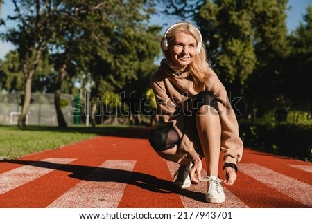 Caucasian mature female runner athlete tying training shoes while jogging on the stadium in public park in the morning. Active healthy sporty lifestyle.