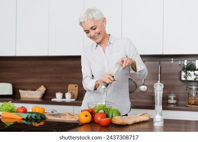 Caucasian mature housewife seasoning with spices salt pepper vegetable salad while cooking at home kitchen. Middle-aged woman preparing dinner lunch. Vegetarianism concept - Powered by Shutterstock