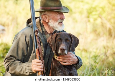 Caucasian Mature Man With Gun And Dog Sit Searching Prey. Bearded Man In Hunting Clothes. Autumn