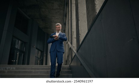 Caucasian mature old man businessman senior business employer walking outdoors on stairs near office building talking phone check time looking at wrist watch late rush hurry worried running lateness - Powered by Shutterstock