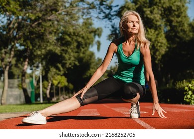 Caucasian mature pretty slim woman in sporty outfit stretching her leg on stadium during yoga class in park outdoors. Slimming training. - Powered by Shutterstock