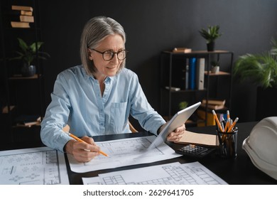 Caucasian mature woman architect drawing blueprints in modern office. Focused woman designer engineer working on new residence complex architectural project using tablet. Construction, engineering