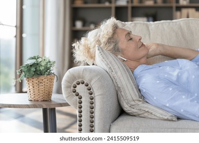 Caucasian mature woman listen music through modern wireless earphones, lying on cozy sofa in living room, chilling alone at home, looks peaceful and quiet, immersed in meditation tune, relaxing indoor - Powered by Shutterstock