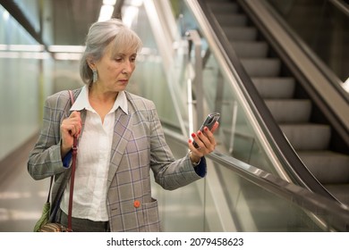 Caucasian Mature Woman Using Her Smartphone While Walking Inside Office Building Near Escalator.