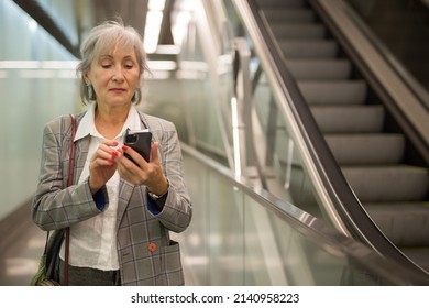 Caucasian Mature Woman Using Her Smartphone While Walking Inside Office Building Near Escalator.