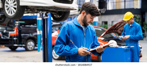 Caucasian Mechanic In Blue Work Wear Uniform Checks The Vehicle Maintenance Checklist With Blur Lifted Car In The Background. Automobile Repairing Service, Professional Occupation.