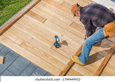 Caucasian Men In His 40s Building Wooden Deck On His Backyard Attaching Wood Board To Each Other Using Cordless Drivers And Screws. Construction Industry Theme.