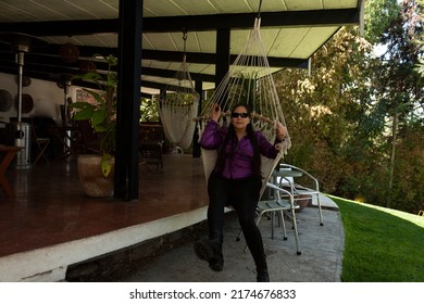 Caucasian Mexican Woman Sitting On A Swing Made Of Fabric Swing Dressed In A Purple Blouse, Pants And Boots With A Happy Face Exercising With Her Legs