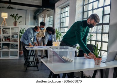 Caucasian Mid Adult Businessman With Face Mask Unpack His Box With Belongings After He Return To Work In The Office While His Coworkers Working Behind