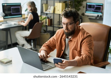Caucasian middle aged man developing neural network using laptop and smartphone at desk, young Caucasian woman coding on computer in background, modern office workspace