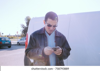 Caucasian Middle Aged Man Wearing A Leather Jacket And Sunglasses Checks His Cell Phone In A Parking Area Before Getting Into His Van