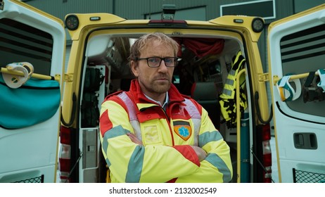 Caucasian Middle Aged Paramedic Standing At The Back Of An Ambulance Car With Confidence Looking Directly At The Camera. Emergency Medical Care Worker Posing Next To The Ambulance Car.	