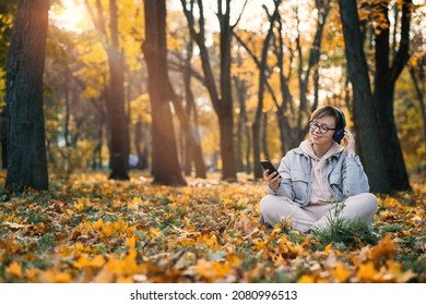 Caucasian Middle Aged Woman In Earphones Listening To Music, Meditation App On Smartphone And Meditating In Lotus Pose At Autumn Park. Meditation App, Mental Health, Self Care, Mindfulness Concept