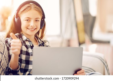 Caucasian middle schoolgirl with laptop pc and headphones at home showing thumb up and smiling. Distance education, free time, technology and internet concept