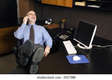Caucasian middle-aged businessman in office sitting with feet on desk talking and laughing on cellphone.