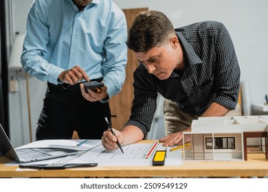 A Caucasian middle-aged male engineer contractor and an Italian architect engineer are seated at a desk, discussing renovation plans with blueprints and a house model in front of them. - Powered by Shutterstock