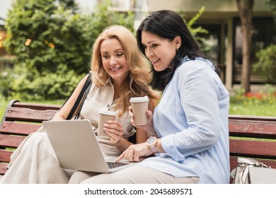 Caucasian Middle-aged Mature Women Sisters Best Friends Girlfriends Watching Movie Webinar Working Online Remotely Together, Using E-banking E-commerce On Computer In City Park Drinking Coffee