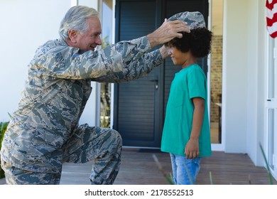 Caucasian Military Grandfather In Camouflage Clothing Wearing Cap To Biracial Grandson Outside House. Unaltered, Family, Togetherness, Childhood, Military, Patriotism And Homecoming.