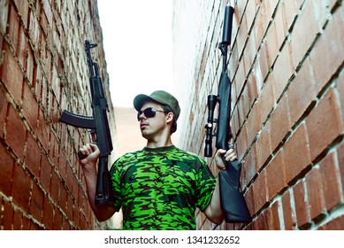 Caucasian Military Man With Black Sunglasses Indoor Urban Room Space Stand With Machine Gun And Rifle With A Telescopic Sight Near Red Brick Wall. Corridor In Perspective.Empty Space For Inscription.