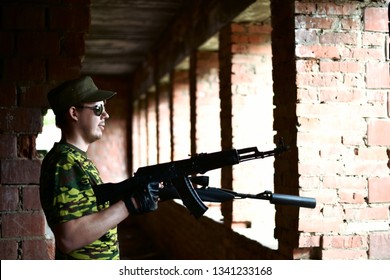 Caucasian Military Man With Black Sunglasses Indoor Urban Room Space Stand With A Kalashnikov Rifle Near Abandoned Red Brick Wall With Windows. Corridor In Perspective. Empty Space For Inscription