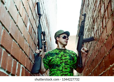 Caucasian Military Man With Black Sunglasses Indoor Urban Room Space Stand With Machine Gun And Rifle With A Telescopic Sight Near Red Brick Wall. Corridor In Perspective.Empty Space For Inscription.