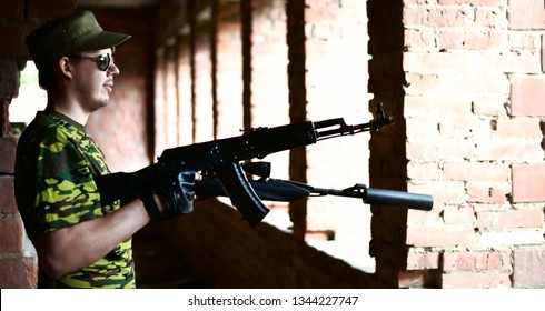 Caucasian Military Man With Black Sunglasses Indoor Urban Room Space Stand With A Kalashnikov Rifle Near Abandoned Red Brick Wall With Windows. Corridor In Perspective. Empty Space For Inscription