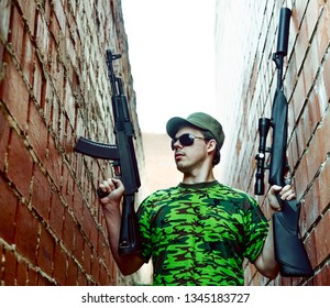 Caucasian Military Man With Black Sunglasses Indoor Urban Room Space Stand With Machine Gun And Rifle With A Telescopic Sight Near Red Brick Wall. Corridor In Perspective.Empty Space For Inscription.