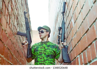 Caucasian Military Man With Black Sunglasses Indoor Urban Room Space Stand With Machine Gun And Rifle With A Telescopic Sight Near Red Brick Wall. Corridor In Perspective.Empty Space For Inscription.